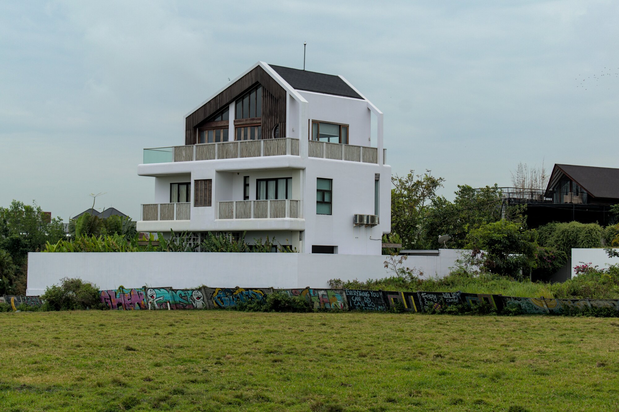 A striking large, white three-story villa behind a grass field with a low fence covered in colorful graffiti, mostly in English.