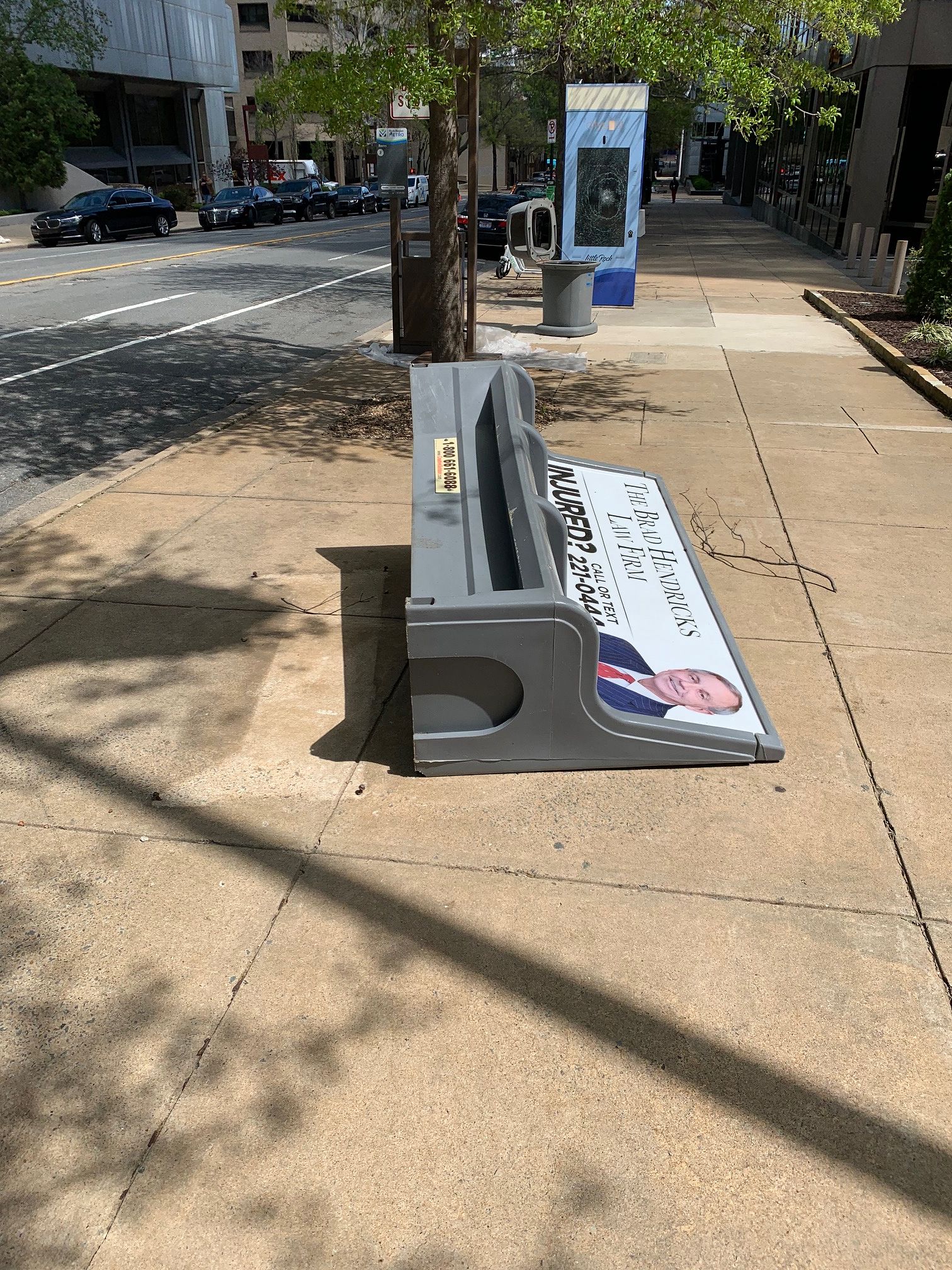 A bus bench with an ad for a personal injury lawyer on its back, unanchored from the pavement and tipped over so the lawyer is sprawled on his back.